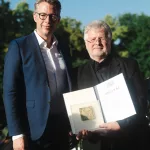 Bernd SChweinar und Staatsminister Markus Blume. Bernd SChweinar hält eine Mappe in der Hand.