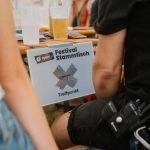 Man blickt zwischen zwei sitzenden Personen durch. Man sieht nur jeweils einen Arm von hinten. Zentral: Biertisch, auf dem ein Bierbecher steht und ein Schild herunterhängt, auf dem zu lesen ist "Vaupebai Festivalstammtisch Treffpunkt".