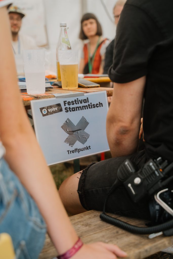 Man blickt zwischen zwei sitzenden Personen durch. Man sieht nur jeweils einen Arm von hinten. Zentral: Biertisch, auf dem ein Bierbecher steht und ein Schild herunterhängt, auf dem zu lesen ist "Vaupebai Festivalstammtisch Treffpunkt".